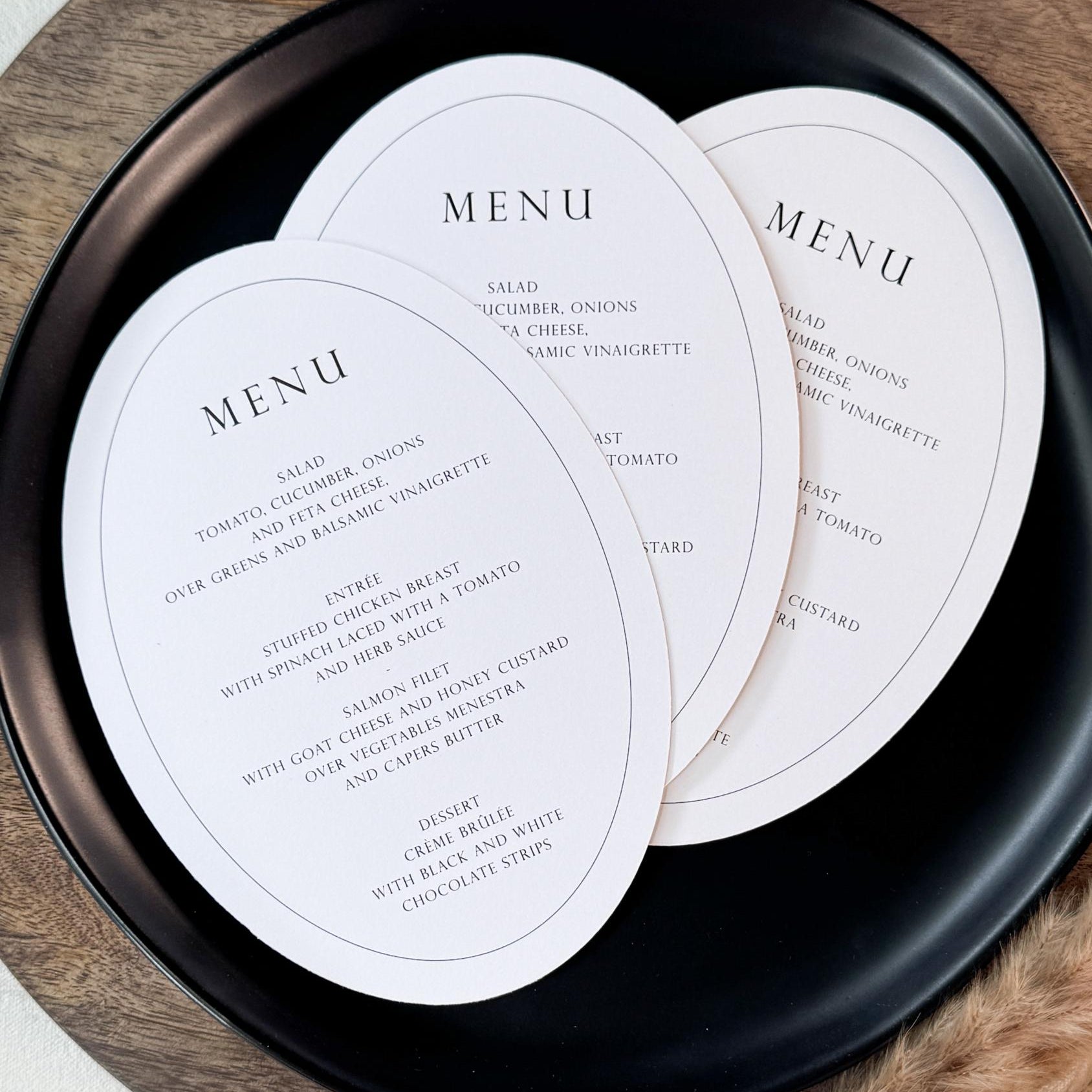 Three oval white menu cards printed with elegant black text, arranged on a round black plate over a wooden board with dried grass stems on a light beige surface.