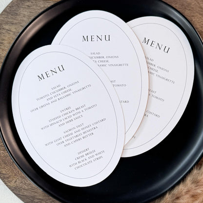 Three oval white menu cards printed with elegant black text, arranged on a round black plate over a wooden board with dried grass stems on a light beige surface.