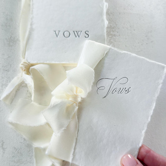 A white paper vows booklet with a cream satin ribbon bow, held by a hand against a textured light gray background. The booklet is labeled 'VOWS' and features elegant script and serif typography.