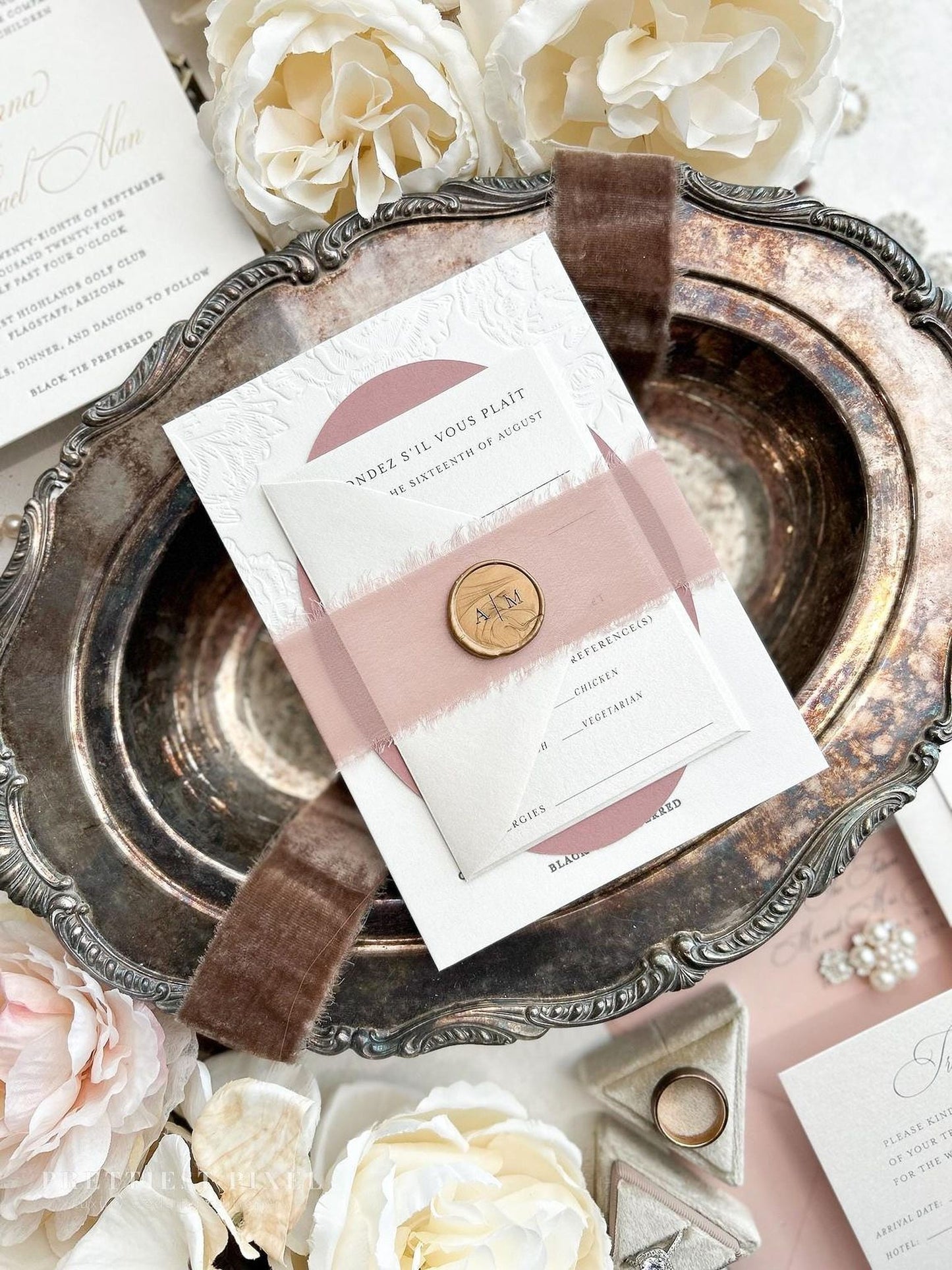 a wedding invitation with a pink and white color scheme, placed on a decorative metal tray. The invitation includes text and a small gold seal, suggesting it is a formal wedding invitation.