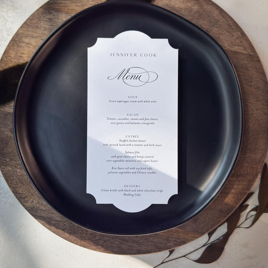 A black ceramic dinner plate with a white menu card placed on top, set on a round wooden charger plate against a light neutral background with soft shadows.