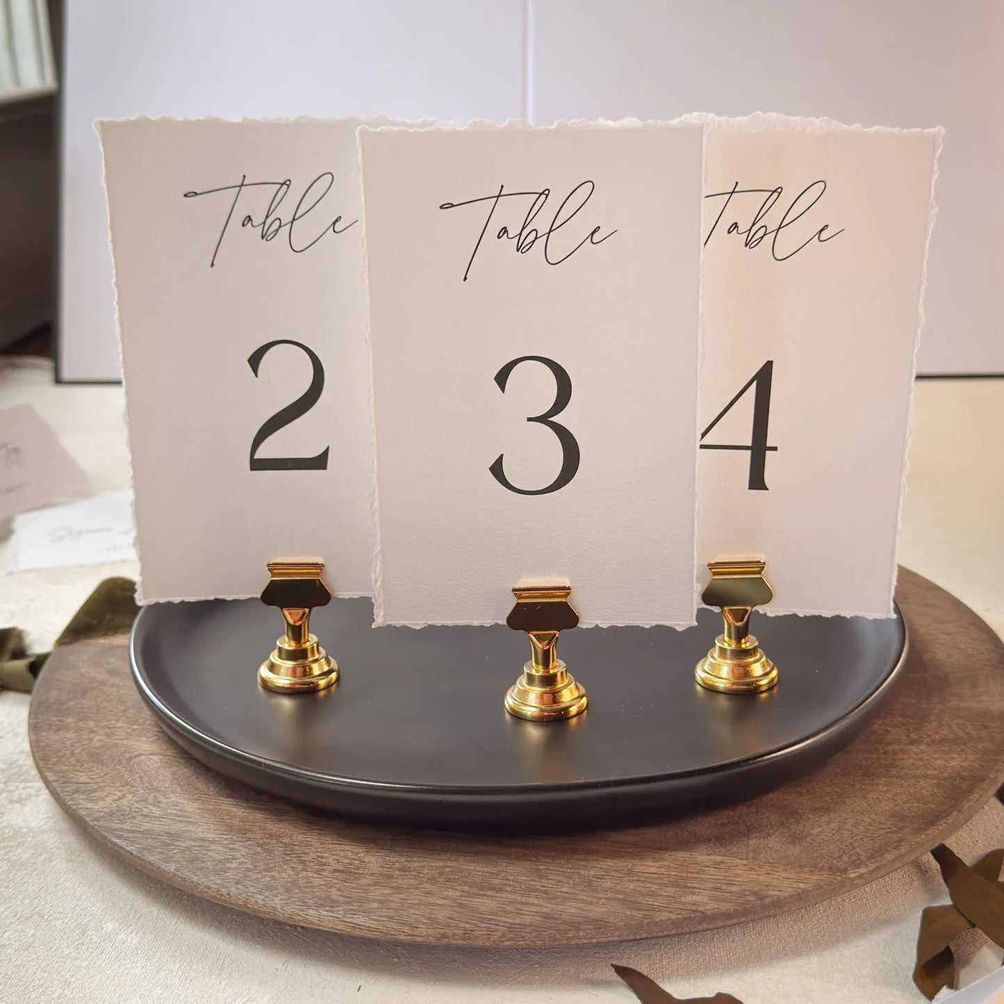 A table number display with three paper cards, each displaying a number and the word "Table" written on it. The cards are attached to a black stand with gold clamps.