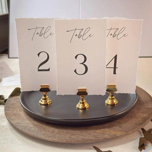 A table number display with three paper cards, each displaying a number and the word "Table" written on it. The cards are attached to a black stand with gold clamps.