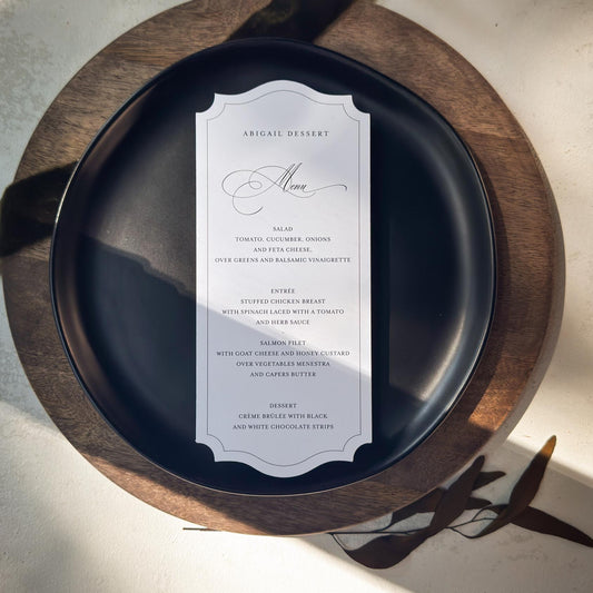 A vertical white wedding invitation card with elegant black script and serif text, styled on a dark navy blue plate over a round wooden charger, with a dark green velvet ribbon on a textured light beige surface.