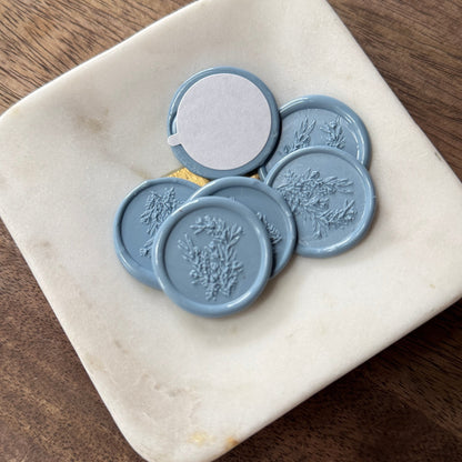 A set of round blue wax seals with embossed floral designs, including one seal showing a white backing, displayed on a small square cream dish over a dark wood surface.