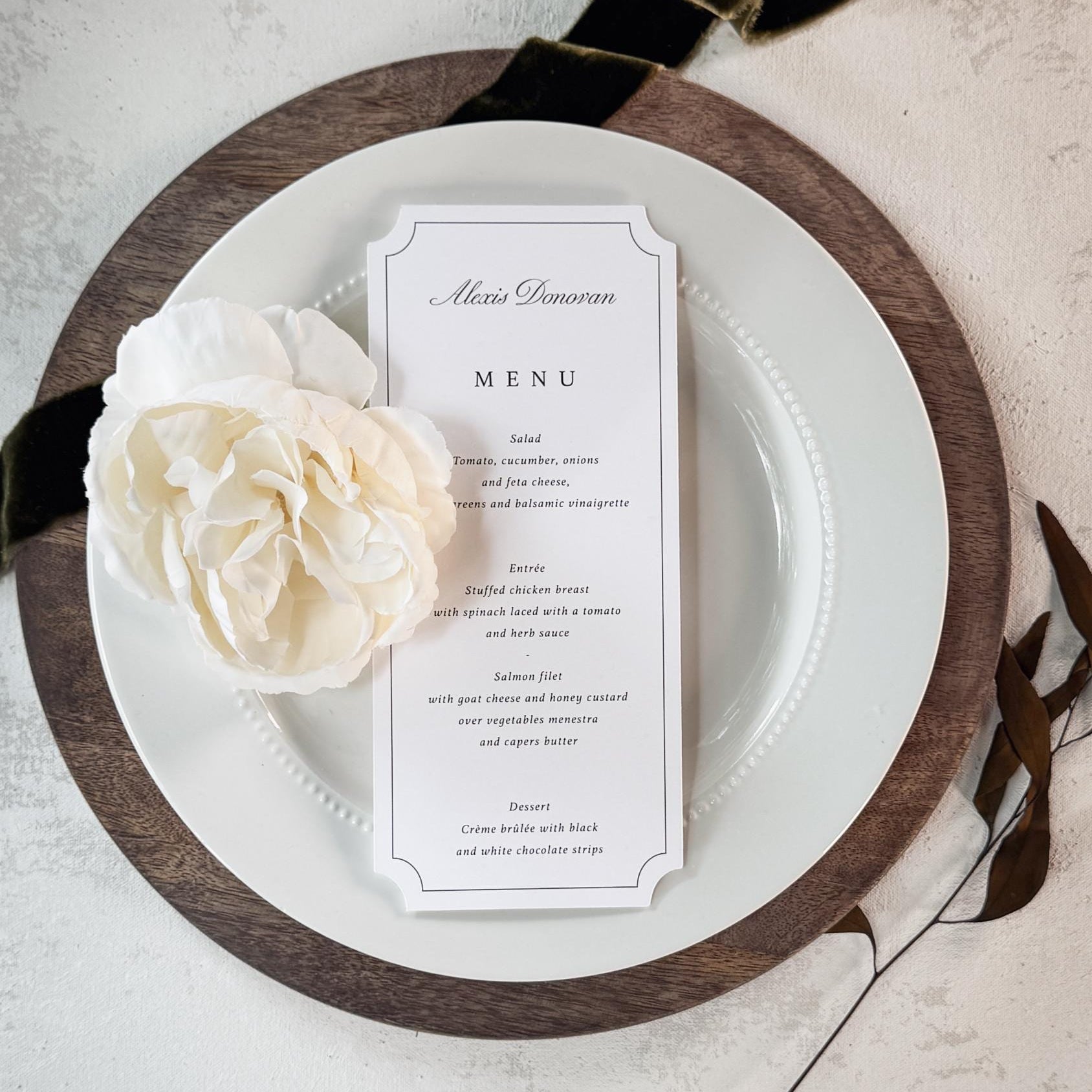 A vertical wedding menu card with elegant black typography on a white card, styled on a white plate with a dark wood charger and a cream flower, set against a textured light gray background with a black ribbon.