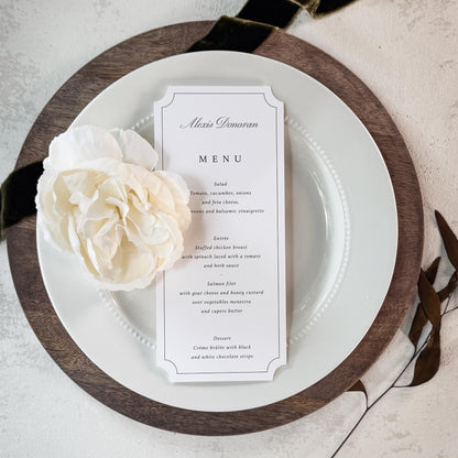 A vertical wedding menu card with elegant black typography on a white card, styled on a white plate with a dark wood charger and a cream flower, set against a textured light gray background with a black ribbon.