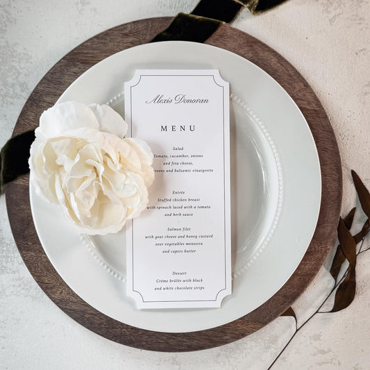 A vertical wedding menu card with elegant black typography on a white card, styled on a white plate with a dark wood charger and a cream flower, set against a textured light gray background with a black ribbon.
