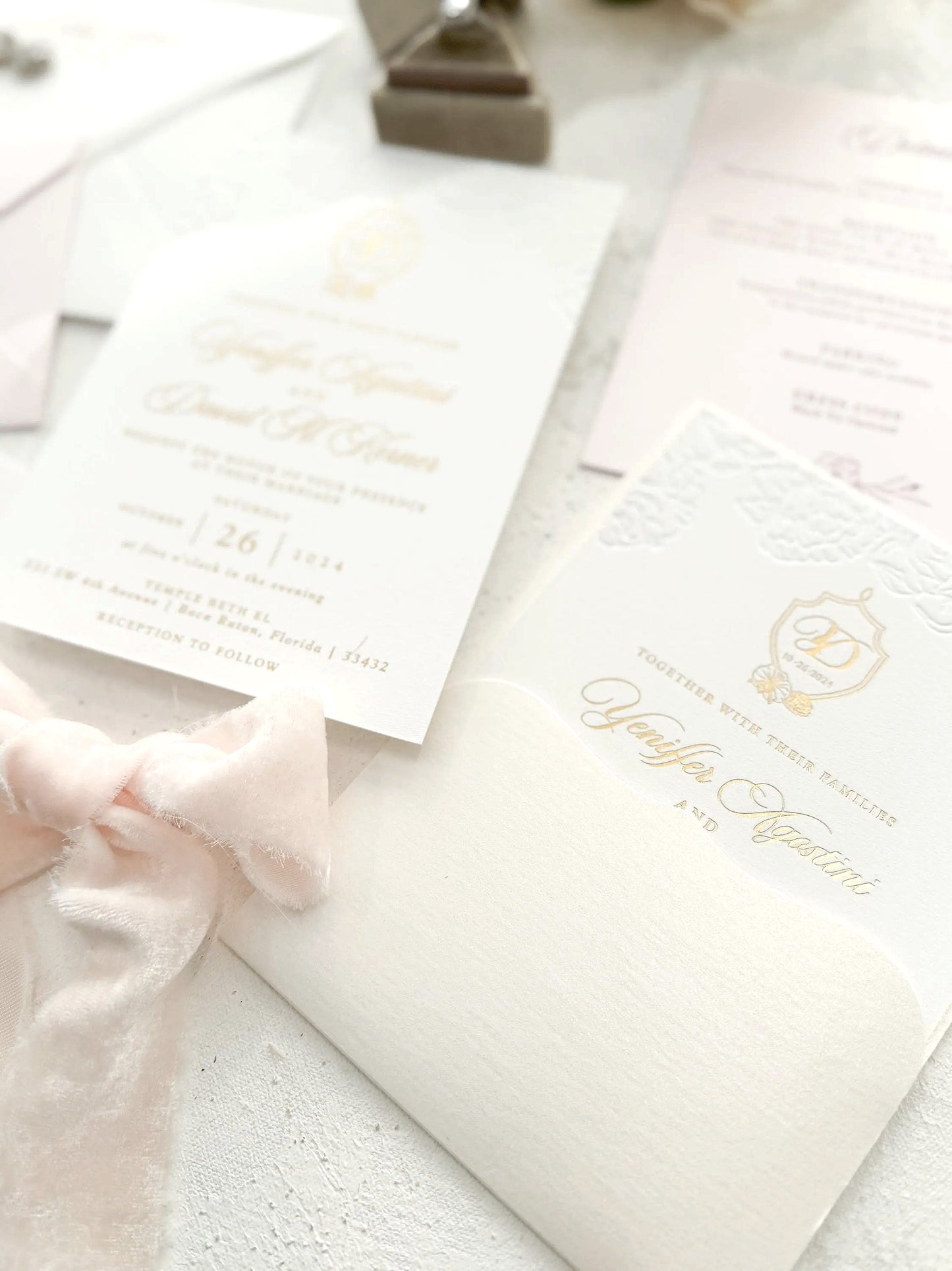 A close-up view of a wedding invitation with a white background and gold lettering, featuring a delicate pink ribbon and a gold emblem.