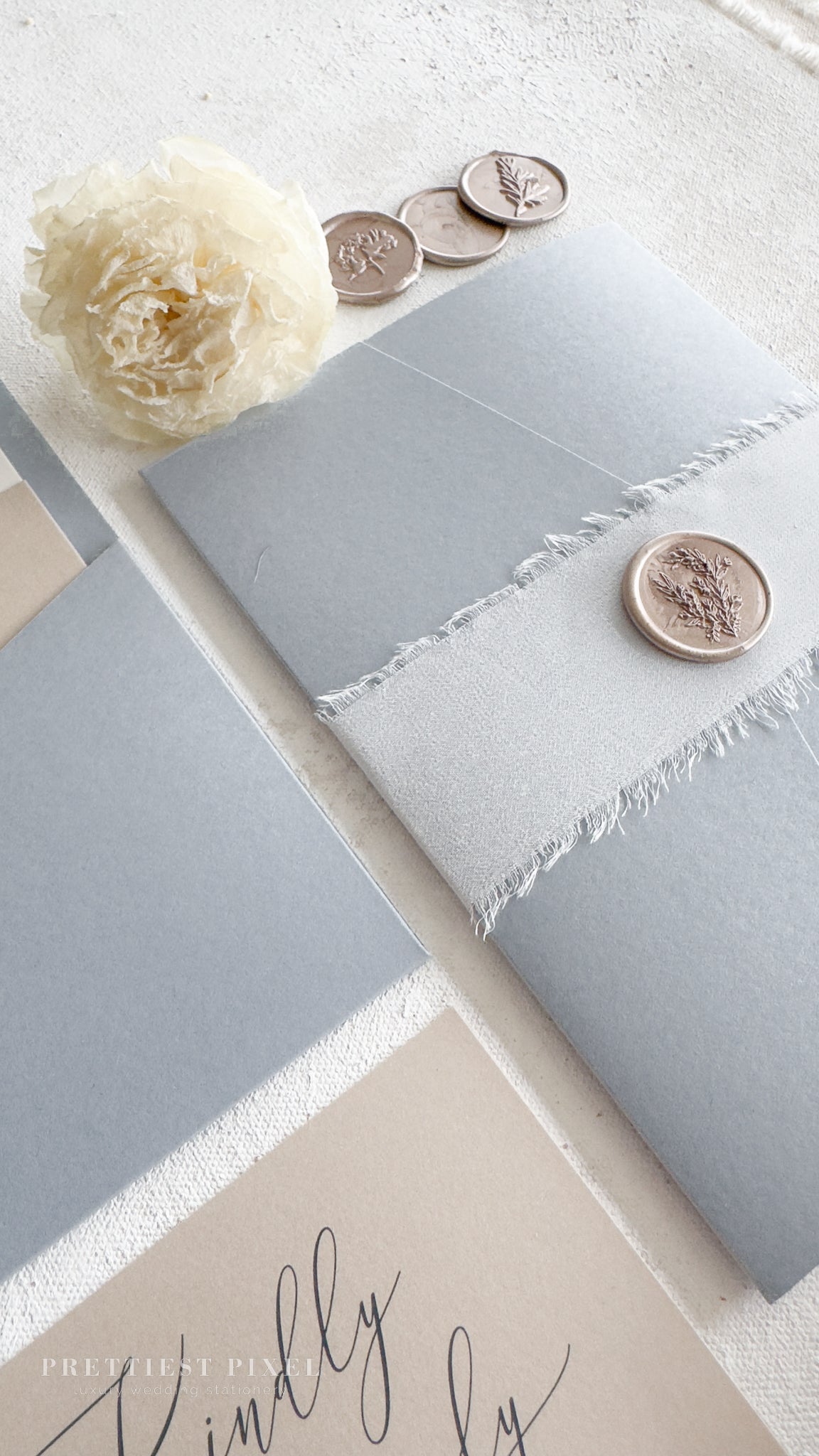 a close-up view of a grey envelope with a white flower and three silver coins with floral designs on them, placed on a white surface.